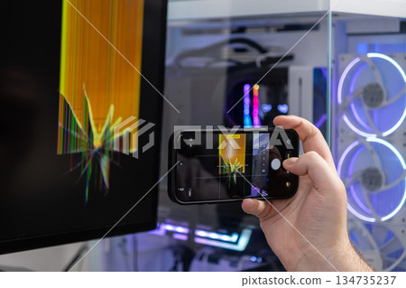 Person's hand using a smartphone to photograph a severely cracked computer monitor screen displaying distorted lines, documenting device malfunction for technical repair 134735237