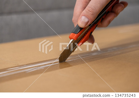 Person's hand skillfully uses a sharp utility knife to carefully cut open a sealed brown cardboard box, symbolizing unboxing and efficient package delivery Person's hand skillfully uses a sharp utility knife to carefully cut open a sealed brown cardboard box, symbolizing unboxing and efficient package delivery 134735243
