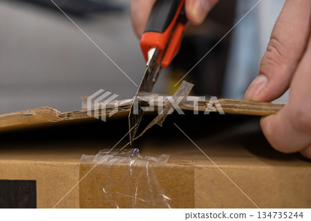 Human hands use a sharp utility knife to carefully cut open a sealed cardboard delivery box, symbolizing e-commerce, logistics, and secure package unpacking 134735244