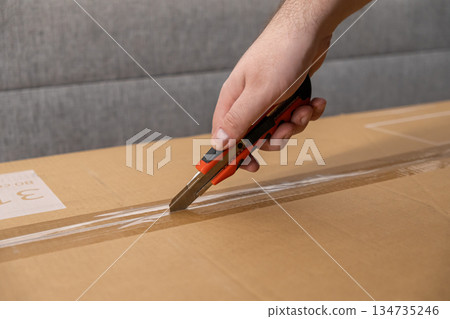Hand using a red box cutter to slice packing tape on a brown cardboard box, symbolizing efficient parcel delivery and quick unpacking process Hand using a red box cutter to slice packing tape on a brown cardboard box, symbolizing efficient parcel delivery and quick unpacking process 134735246