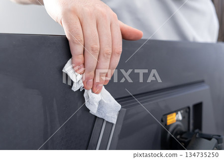 Hand diligently cleaning the dusty back panel of a computer monitor with a wet wipe, emphasizing workstation hygiene and equipment maintenance 134735250