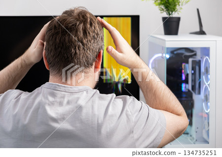 Frustrated man clutching head, gazing at a desktop computer monitor showing severe glitch errors, symbolizing technical malfunction and urgent repair need 134735251