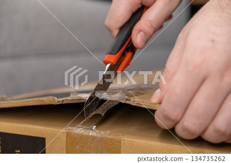 Human hands cutting open a taped brown cardboard shipping box with a sharp utility knife, representing unboxing an online purchase or package delivery Human hands cutting open a taped brown cardboard shipping box with a sharp utility knife, representing unboxing an online purchase or package delivery 134735262