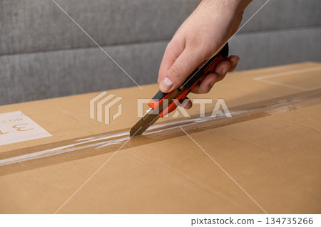 Human hand precisely using a sharp utility knife to open a sealed brown cardboard box, representing e-commerce delivery and customer unpacking process Human hand precisely using a sharp utility knife to open a sealed brown cardboard box, representing e-commerce delivery and customer unpacking process 134735266