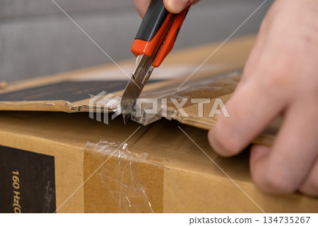 Person's hand skillfully uses a utility knife to open a brown cardboard delivery box, revealing 160 Hz, symbolizing new product unboxing and online retail Person's hand skillfully uses a utility knife to open a brown cardboard delivery box, revealing 160 Hz, symbolizing new product unboxing and online retail 134735267