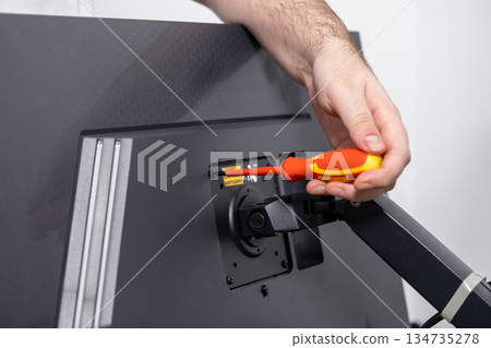 Person's hand uses a precision screwdriver to securely fasten a monitor arm onto a computer display, illustrating practical home office equipment assembly and installation Person's hand uses a precision screwdriver to securely fasten a monitor arm onto a computer display, illustrating practical home office equipment assembly and installation 134735278