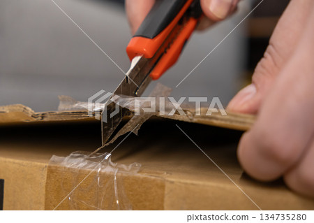 Hand carefully cutting clear tape on a brown cardboard box with a sharp utility knife, symbolizing efficient package opening and delivery 134735280