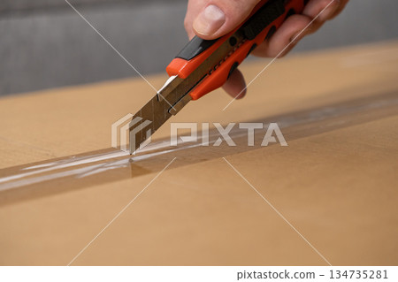 Hand carefully using a sharp orange utility knife to open a brown cardboard delivery box sealed with clear packing tape, illustrating safe unboxing Hand carefully using a sharp orange utility knife to open a brown cardboard delivery box sealed with clear packing tape, illustrating safe unboxing 134735281