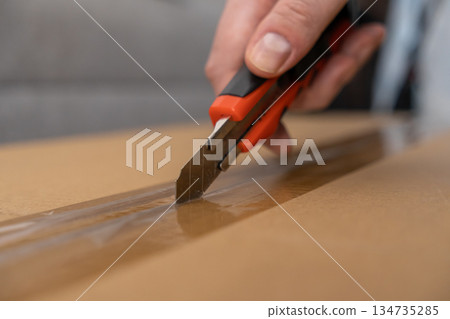 Person's hand with a sharp red utility knife expertly slicing transparent packaging tape on a brown cardboard box, symbolizing efficient home delivery service 134735285