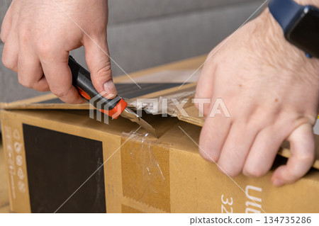 Person's hands using a sharp utility knife to open a sealed brown cardboard box, symbolizing online shopping, home delivery, and consumer purchase unpacking 134735286