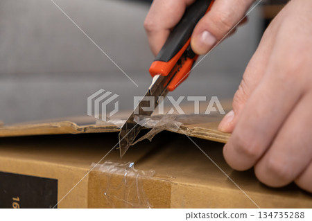 Person carefully opening a new brown cardboard shipping package with a sharp utility knife, symbolizing efficient unboxing, e-commerce delivery, and convenient parcel reception 134735288