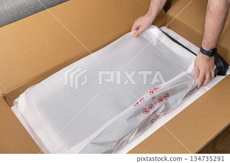 Consumer carefully unpacks new electronic display monitor from a large brown delivery box, signifying modern online retail and purchase fulfillment Consumer carefully unpacks new electronic display monitor from a large brown delivery box, signifying modern online retail and purchase fulfillment 134735291