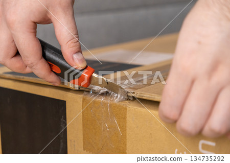 Hand uses utility knife to cut clear tape on brown cardboard delivery box, symbolizing modern e-commerce and new product unpacking 134735292
