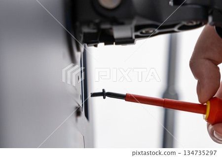 Technician's hand precisely tightens a black screw using an insulated red screwdriver on a wall mount, emphasizing professional assembly and careful maintenance service Technician's hand precisely tightens a black screw using an insulated red screwdriver on a wall mount, emphasizing professional assembly and careful maintenance service 134735297