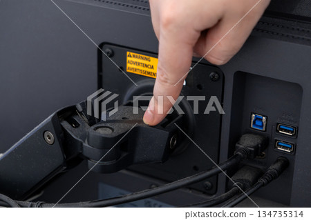Hand adjusting a VESA monitor arm mount on a computer screen, visible warning label and cables, demonstrating ergonomic desk setup 134735314