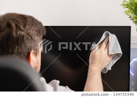 Person cleaning a dusty black computer monitor screen with a grey microfiber cloth at a modern home office desk, emphasizing hygiene and maintenance Person cleaning a dusty black computer monitor screen with a grey microfiber cloth at a modern home office desk, emphasizing hygiene and maintenance 134735340
