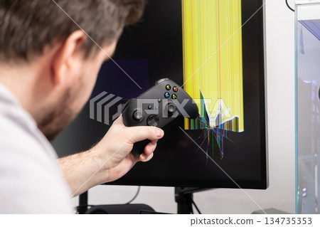 Frustrated gamer holding controller observing a damaged monitor with a severely cracked screen displaying colorful pixels, illustrating intense gaming frustration Frustrated gamer holding controller observing a damaged monitor with a severely cracked screen displaying colorful pixels, illustrating intense gaming frustration 134735353