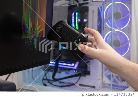 Young gamer's hand holds a dark wireless video game controller against a severely cracked computer monitor, displaying broken colorful pixels, symbolizing intense gamer frustration Young gamer's hand holds a dark wireless video game controller against a severely cracked computer monitor, displaying broken colorful pixels, symbolizing intense gamer frustration 134735354
