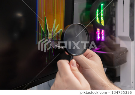 Person meticulously uses a magnifying glass to inspect a severely cracked computer monitor screen displaying vertical lines, representing technology repair and device malfunction 134735356