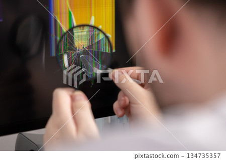 Person using a magnifying glass to closely examine a severely cracked and glitching computer screen, symbolizing electronic device damage and the need for repair service 134735357