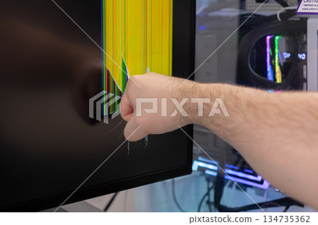 Frustrated individual's angry fist impacts a computer monitor, completely shattering the screen with vibrant, damaged pixel lines, representing digital breakdown and technical rage 134735362