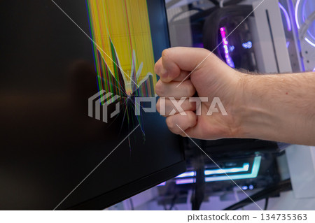 Man's clenched fist near a severely shattered computer monitor displaying colorful broken pixels, symbolizing extreme frustration and technology failure 134735363