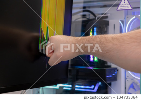 Aggressive persons fisted hand violently hits a gaming monitor, shattering the screen into a vibrant display of broken pixels, portraying intense digital frustration and tech rage 134735364