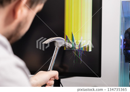 An angry person holds a hammer aiming at a shattered computer monitor displaying vibrant broken pixel lines, symbolizing overwhelming technological frustration or system failure 134735365