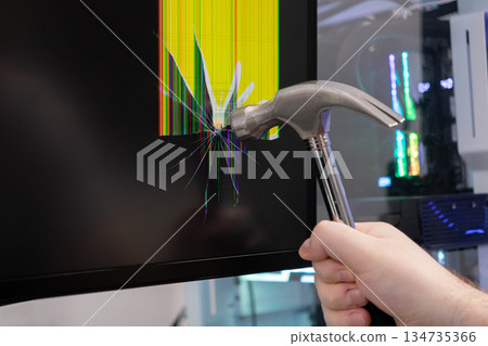 Hand with a hammer violently smashes a computer monitor, creating a shattered screen with colorful pixel lines, symbolizing technology frustration, burnout, or system failure 134735366