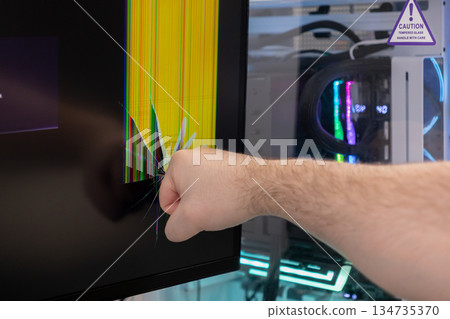 A person's fist strikes a computer monitor, causing a badly cracked screen with colorful vertical lines, conveying intense anger, technology frustration, and system failure 134735370