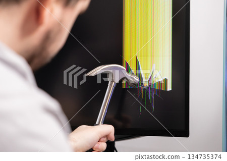 Angry individual smashes a computer monitor screen with a hammer, causing severe cracks and vibrant lines, symbolizing technology frustration 134735374