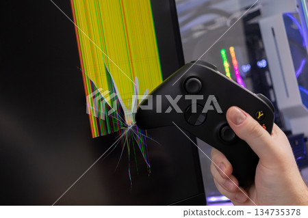 Hand holding a black game controller smashing into a computer monitor, causing widespread screen cracks and illustrating intense gaming frustration 134735378