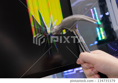 Hand with hammer smashes computer monitor, causing shattered screen with digital distortion and broken pixels, representing technological frustration and system error 134735379