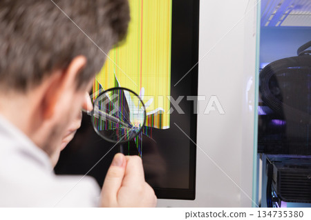 Expert technician uses a magnifying glass to inspect a cracked computer monitor screen, depicting electronic device repair, damage assessment, and IT support 134735380