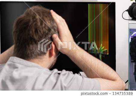 Man reacting with frustration to a severely broken computer monitor displaying vibrant vertical lines and a visible crack, symbolizing sudden technological failure Man reacting with frustration to a severely broken computer monitor displaying vibrant vertical lines and a visible crack, symbolizing sudden technological failure 134735389