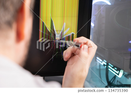A technician points a screwdriver at a severely cracked computer monitor displaying colorful vertical lines, symbolizing urgent hardware failure and tech repair needs 134735390