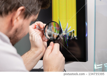 Technician examines cracked computer monitor with magnifying glass and screwdriver, symbolizing electronics repair and technical diagnostics 134735395