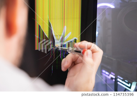 Hand with a screwdriver touching a cracked monitor screen, displaying colorful dead pixels, symbolizing electronic device malfunction and urgent repair 134735396