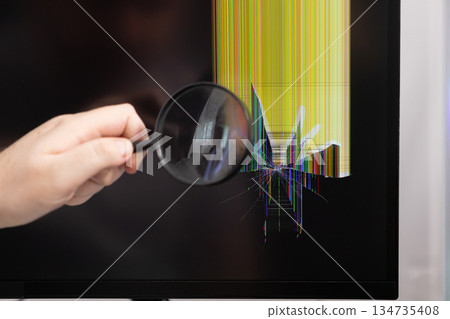 Hand holding magnifying glass inspecting a severely cracked computer monitor screen with colorful malfunction lines, symbolizing technical problem diagnosis 134735408