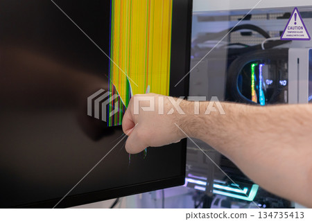 Man's aggressive fist strikes a modern computer monitor screen, causing severe cracking, pixel distortion, and colorful vertical lines, representing frustration or damage Man's aggressive fist strikes a modern computer monitor screen, causing severe cracking, pixel distortion, and colorful vertical lines, representing frustration or damage 134735413