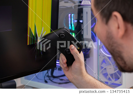 Angry gamer holds black controller near severely cracked monitor displaying corrupted lines, depicting gaming rage, stress, and technology damage Angry gamer holds black controller near severely cracked monitor displaying corrupted lines, depicting gaming rage, stress, and technology damage 134735415
