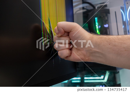 Male clenched fist smashes computer monitor, causing shattered screen with colorful pixel lines, symbolizing extreme frustration and tech rage 134735417