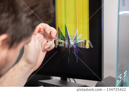 Hand holds screwdriver, pointing at computer monitor with severely cracked screen displaying colorful glitch lines, representing technical malfunction, repair needs Hand holds screwdriver, pointing at computer monitor with severely cracked screen displaying colorful glitch lines, representing technical malfunction, repair needs 134735422
