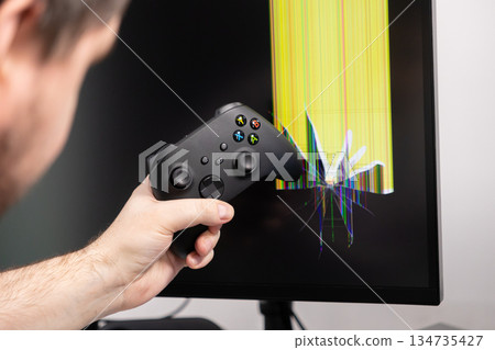 A frustrated gamer's hand holds a black controller, aggressively positioned against a broken computer monitor with a shattered screen and bright pixel lines 134735427