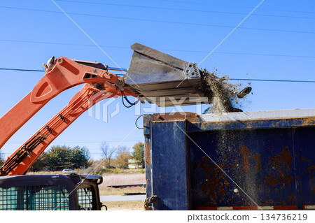 Excavator machine with large bucket pours waste dirt into container at construction site. Excavator machine with large bucket pours waste dirt into container at construction site. 134736219