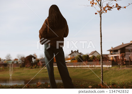 Woman in casual attire looking at a scenic view of a lake and suburban houses. A girl stands with her back to the camera The autumn scenery of a park Woman in casual attire looking at a scenic view of a lake and suburban houses. A girl stands with her back to the camera The autumn scenery of a park 134742832