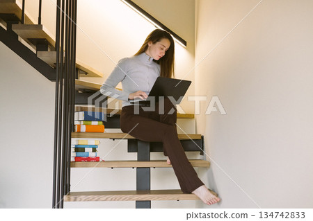 Young woman working on laptop at home office sitting on wooden stairs, cozy minimalist interior, remote work Calm productivity and slow living vibe. 134742833