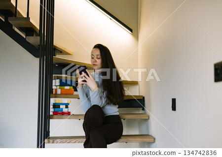 Young brunette woman using smartphone while sitting on modern wooden stairs at home, cozy minimalist interior. Modern female lifestyle. Homy scene. 134742836