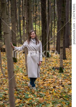Young woman in a beige coat stands among tall trees in an autumn forest, touching a tree trunk, surrounded by yellow leaves and soft natural light Young woman in a beige coat stands among tall trees in an autumn forest, touching a tree trunk, surrounded by yellow leaves and soft natural light 134743891