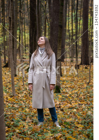 Woman in a light beige coat stands in an autumn forest, looking up among tall trees and yellow fallen leaves, calm and reflective mood 134743892